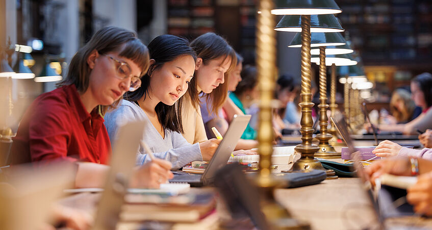 Mehrere Studierende sitzen im Großen Lesesaal der Universität Wien und lernen bei Lampeschein mit Laptops und Büchern.